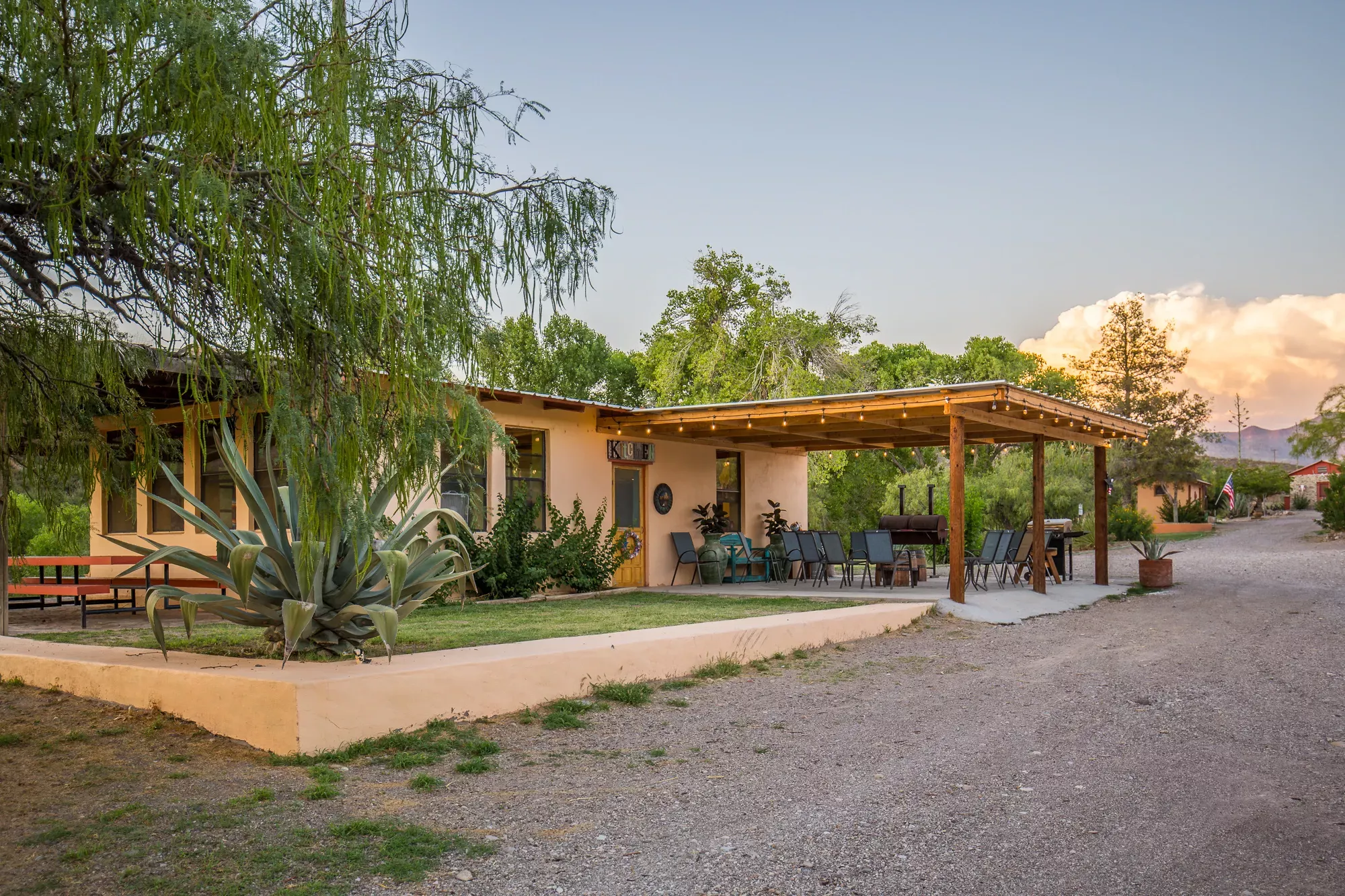 A light orange/pink casita with string lights along the awning with tables and chairs, as well as a barbeque pit are central, with small casitas in the background.